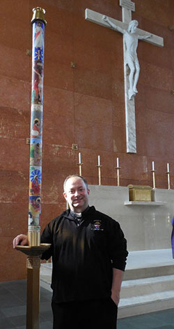 Father Andrew Budzinski, pastor of St. John the Baptist Parish, Fort Wayne, stands by the Paschal Candle decorated by parishioner Art Cislo.