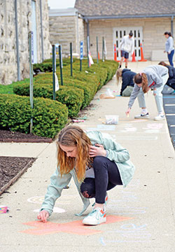 Bailey Freese and other members of the eighth-grade class of St. Luke School in Indianapolis decorate the sidewalk around the parish church on April 5, Palm Sunday. The school’s eighth-grade class traditionally presents a Passion play for the parish and school during Holy Week. The event was not possible this year due to safe-distance guidelines during the coronavirus outbreak. So the classmates—remaining six or more feet apart—participated in a chalk art project on the sidewalks around the church on Palm Sunday morning, writing Scripture passages, encouraging messages and pictures. Parishioners and school families were invited on Palm Sunday afternoon to view the art as well as outdoor Stations of the Cross by car or by walking while maintaining safe distance. An e-mail about the project sent by the Eighth-Grade Room Parent Team to class parents stated, “This is a great way for the Class of 2020 to do our part to help our parish members to connect with St. Luke until we are able to worship together again.” (Photo by Natalie Hoefer) 