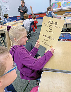 Riley Keal looks at a certificate in Japanese script as Cameron Tipton answers questions about Japan in the background during his visit to the sixth-grade class of his alma mater, St. Gabriel School in Connersville, on Jan. 10. Tipton teaches English as a second language in Japan and helped coordinate a video exchange between his Japanese sixth-grade students and the sixth-graders at St. Gabriel. (Submitted photo by Susie Tipton)