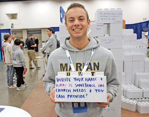 Aaron Bauman, a senior in the San Damiano Scholars program at Marian University in Indianapolis, shows the giant, plastic building block that features an intriguing question for youths at the interactive exhibit hall of the National Catholic Youth Conference in Indianapolis on Nov. 20. (Photo by John Shaughnessy)