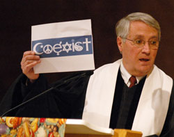 Dr. Robert Welsh, the president of the Council on Christian Unity of the Indianapolis-based Christian Church (Disciples of Christ), holds up an image of a bumper sticker which uses religious symbols to spell the word “coexist” to invite those in attendance to consider that the various world faith traditions are called to more than coexistence, but also to build community. (Photo by Sean Gallagher)