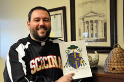 Father Aaron Jenkins holds a copy of the coat of arms that he designed for Bishop-designate Christopher J. Coyne. Father Jenkins is the associate director of vocations for the archdiocese, chaplain of Father Thomas Scecina Memorial High School in Indianapolis, chaplain of the Indianapolis Fire Department, and sacramental minister of St. Maurice Parish in Napoleon, Immaculate Conception Parish in Milhousen and St. Denis Parish in Jennings County. (Photo by Mary Ann Wyand)