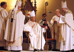Bishop Paul D. Etienne, center, sits in the bishop’s chair, also known as the cathedra, of the Diocese of Cheyenne, Wyo., on Dec. 9, 2009, at the Cheyenne Civic Center during his ordination and installation as the shepherd of the Church in Wyoming. The principal action of installing a bishop as the spiritual leader of a diocese is the seating of him in the cathedra. Applauding Bishop Etienne are, from left, Archbishop Charles J. Chaput, then of Denver, and Archbishop Pietro Sambi, then apostolic nuncio to the United States. Bishop Etienne was previously a priest of the Archdiocese of Indianapolis. (File photo by Sean Gallagher)