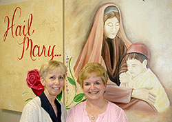 Connie Hessler, left, and Donna Ross are best friends from college who have been teaching fourth-grade classes at St. Jude School in Indianapolis for 40 years. As they prepare to retire on May 23, they pose in front of a mural of the Blessed Mother and Jesus as a boy near their classrooms. The two teachers had the mural created to honor their moms. (Photo by John Shaughnessy)