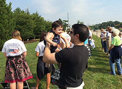 A man lifts a baby while Crossroads walkers pray in the background in front of Planned Parenthood in Indianapolis on July 26. The young adult Crossroad walkers journeyed on foot from San Francisco to Washington, D.C., this summer in support of respect for life. In the archdiocese, they walked along U.S. 40 across the state, stopping in Terre Haute, Indianapolis and Richmond. (Submitted photo)