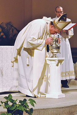 Archbishop Joseph W. Tobin ritually breathes upon chrism oil while blessing it on March 22 during the annual archdiocesan chrism Mass at SS. Peter and Paul Cathedral in Indianapolis. Seminarian Timothy DeCrane, right, assists as an altar server during the liturgy. Oils used in sacraments and the consecration of altars and churches are blessed during the liturgy, which also includes priests renewing their ordination promises. (Photo by Sean Gallagher)