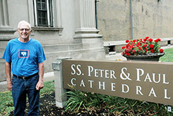 John Meisberger poses for a photo outside SS. Peter and Paul Cathedral in Indianapolis, which was one of the churches that the 72-year-old southern Indiana farmer visited in his successful faith journey to attend Mass at every parish in the archdiocese. (Photo by John Shaughnessy)