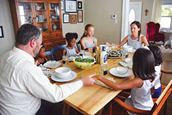 Josh Bach leads his family in a prayer before the meal in their home in Indianapolis on July 10. He and his wife, Cara, members of St. Joan of Arc Parish in Indianapolis, have adopted the three girls seated in the chairs—Amelia, left, and Frances, both 6, and Victoria, 12. They hope to finalize soon the adoption of the two girls seated on the bench, who for legal reasons must remain anonymous until their adoption is complete. (Photo by Natalie Hoefer)