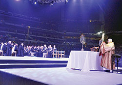 Father Joseph Espaillat II blesses thousands in Lucas Oil Stadium in Indianapolis on Nov. 17 in solemn Benediction during a general session of the National Catholic Youth Conference held in Indianapolis on Nov. 16-18. Kneeling in prayer on the stage during the liturgy are monks of Saint Meinrad Archabbey in St. Meinrad and youth participants in its One Bread One Cup youth liturgical leadership program. They joined more than 20,000 other conference participants in prayer. (Photo by Sean Gallagher)