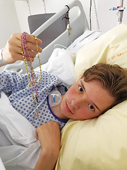 From his hospital bed in France, Alex Kalscheur, 18 and a member of Our Lady of the Greenwood Parish in Greenwood, holds two rosaries he touched to a relic at Notre Dame Cathedral in Paris on July 6. They were still in his pocket later the same day when he fell 30 feet, fracturing one vertebrae and destroying another. (Submitted photo)