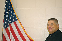 Father Juan Valdes, the administrator of St. Anthony Parish in Indianapolis and a new American citizen, poses for a photo with the American flag inside the gymnasium of the parish school. (Photo by John Shaughnessy)