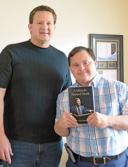 Greg, left, and Mark Hublar pose in Mark’s New Albany apartment with the book Greg wrote about the against-the-odds accomplishments and impact of his brother, who overcame challenges of Down syndrome to become a motivational speaker advocating around the state and nation for the employment of those with disabilities. (Photo by Natalie Hoefer)