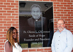 The smiling image of the late Father Glenn O’Connor greets people near the entrance of the new Seeds of Hope residence center on the grounds of St. Joseph Parish in Indianapolis. The archdiocesan priest started the Seeds of Hope program in 1999 to help women overcome their struggles with drugs and alcohol. Standing near the tribute to Father O’Connor are Marvetta Grimes, the executive director of Seeds of Hope, and Sean O’Connor, one of the priest’s seven siblings. (Photo by John Shaughnessy)