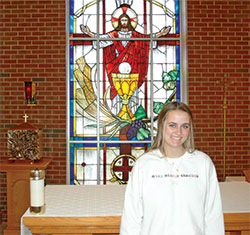 Wearing her sweatshirt stitched with the phrase, “Make Heaven Crowded,” 18-year-old Celia Boring has made it a goal during her senior year at Roncalli High School in Indianapolis to spend more time in eucharistic adoration—and invite others to do the same. Here, she poses in front of a stained-glass window featuring Jesus and the Eucharist in the school’s chapel. (Photo by John Shaughnessy)